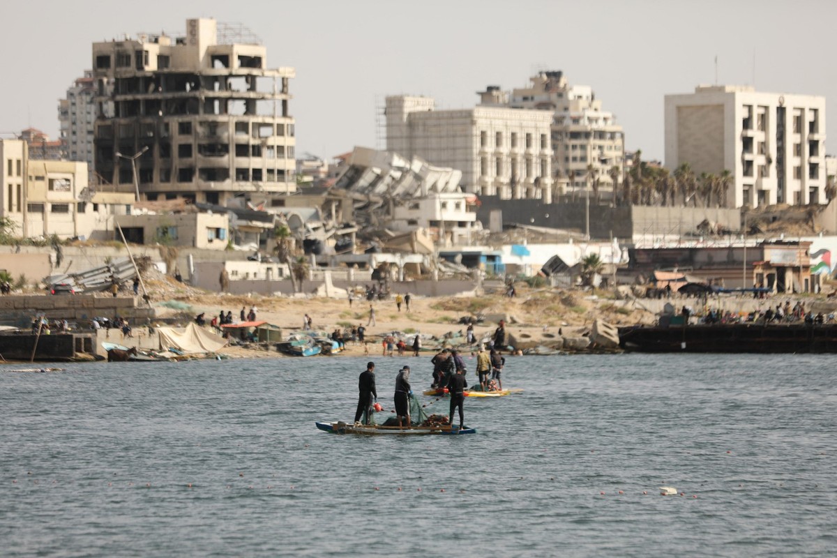 Pescadores palestinos tentam pescar com barcos a remo e varas de pescar na costa da Cidade de Gaza, enquanto prédios danificados e destruídos são vistos ao fundo (Foto: DAWOUD ABO ALKAS / AFP).