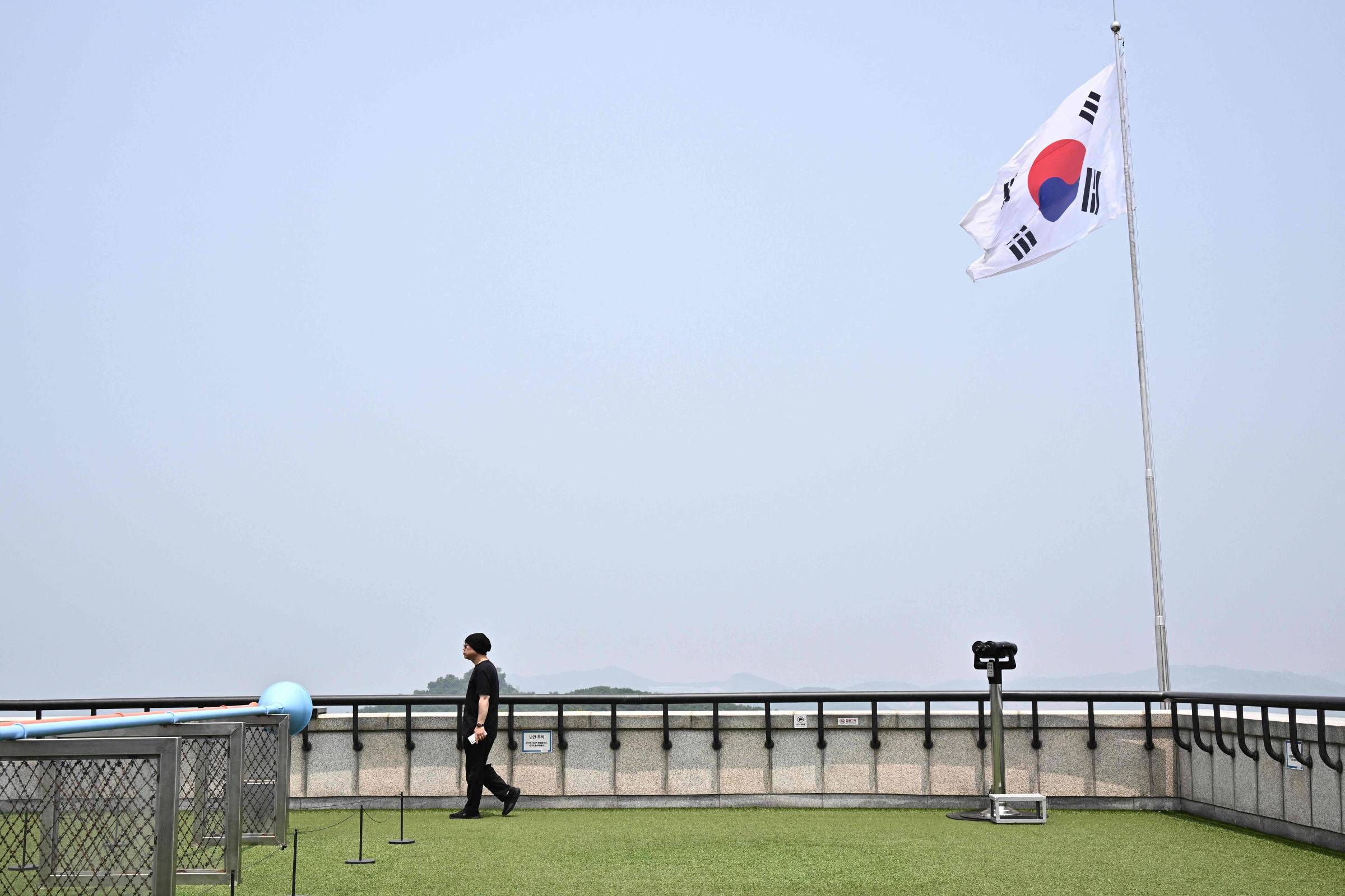 Coreia do Sul inicia desativação de alto-falantes na fronteira com a Coreia do Norte Homem caminha sob a bandeira da Coreia do Sul em um mirante de onde os visitantes podem olhar para a Coreia do Norte (Foto: Anthony Wallace - 11.jun.24/AFP)
