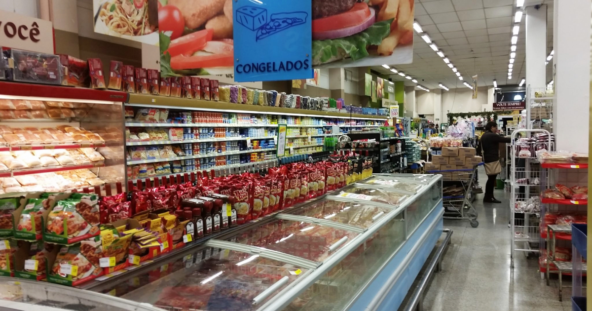 Brasileiros preferem supermercados a feiras livres e isso gera preocupações econômicas Corredor de supermercado com cartazes mostrando alimentos, prateleiras repletas de alimentos processados e embalados e uma geladeira horizontal com produtos congelados (Foto: USP Imagens)
