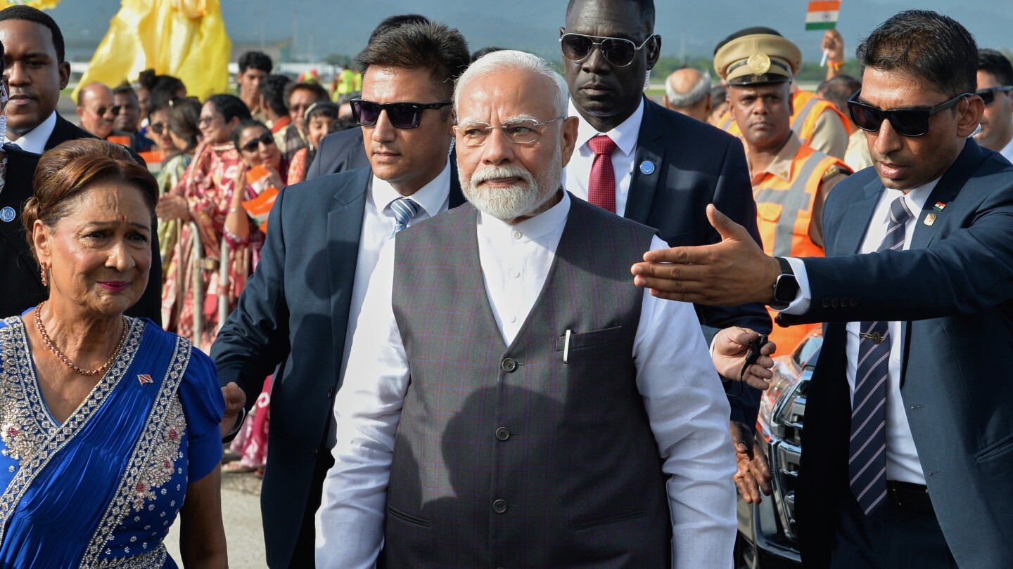 O primeiro-ministro da Índia, Narendra Modi, ao centro, caminha com a primeira-ministra de Trinidad e Tobago, Kamla Persad-Bissessar, vestida com trajes tradicionais indianos, à esquerda, ao chegar no Aeroporto Internacional Piarco em Piarco, Trinidad e Tobago, na quinta-feira, 3 de julho de 2025. (Foto: AP Photo/Jermaine Cruickshank)