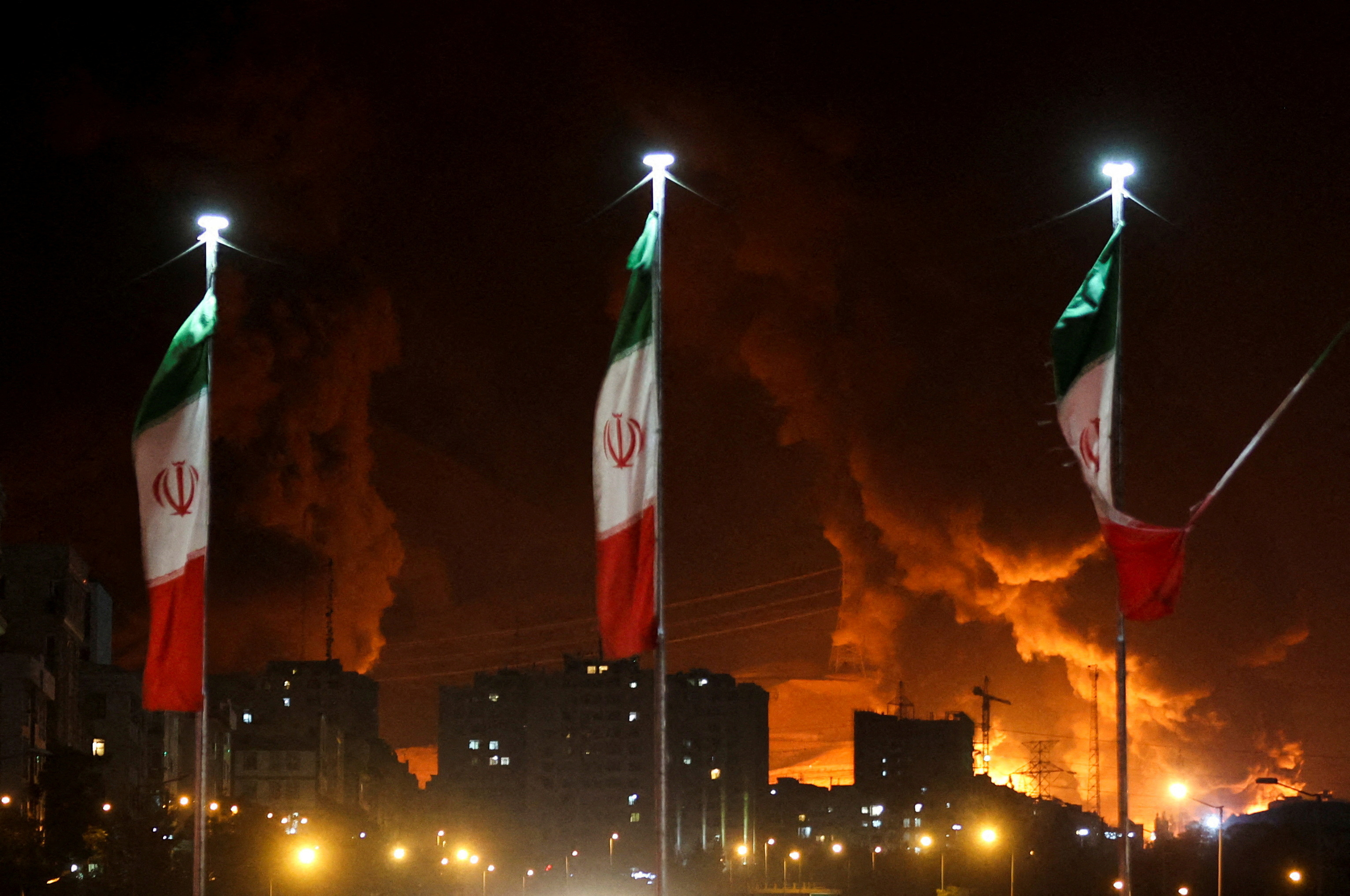 Bandeiras iranianas tremulam enquanto fogo e fumaça de um ataque israelense ao depósito de petróleo de Sharan aumentam, após ataques israelenses ao Irã, em Teerã, Irã, 15 de junho de 2025. (Foto: Majid Asgaripour/WANA (West Asia News Agency) via REUTERS)