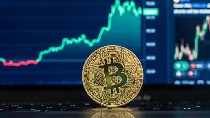 Bitcoin atinge novo recorde histórico acima de US$ 112 mil com alta tecnológica O logotipo da criptomoeda Bitcoin pode ser visto em uma moeda em frente a um gráfico do Bitcoin. (Foto: Silas Stein | Picture Alliance | Getty Images)